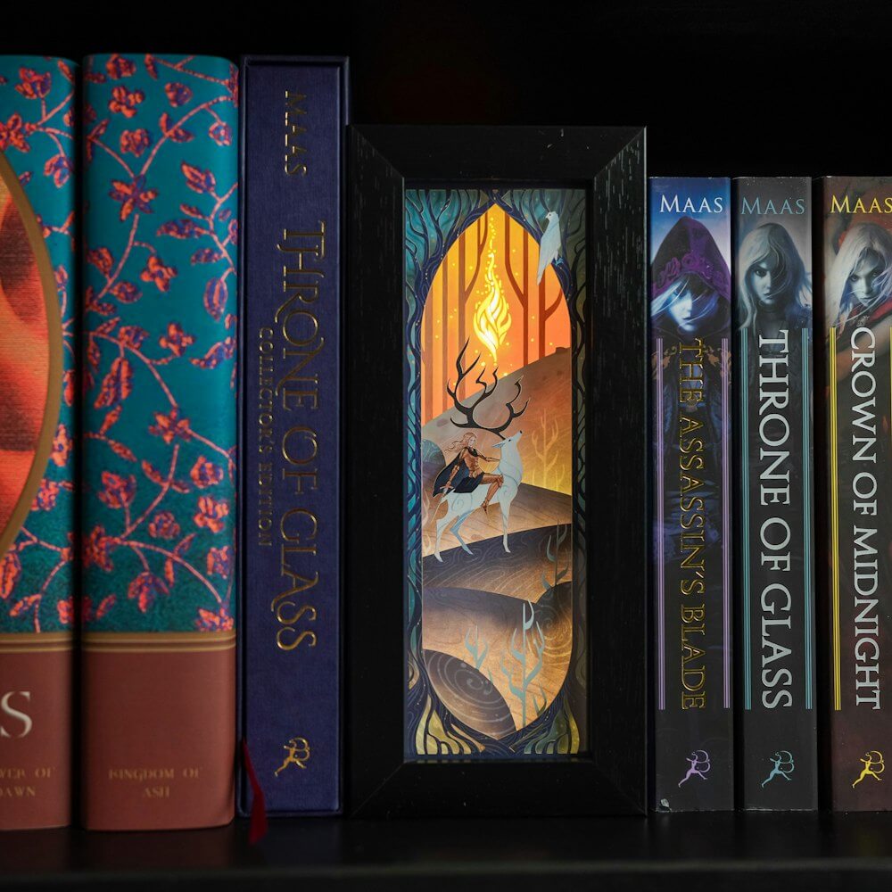 Purple, blue, and yellow Throne of Glass Bookshelf Alley is a black-framed shadowbox with a white papercut design of a woman on a stag.