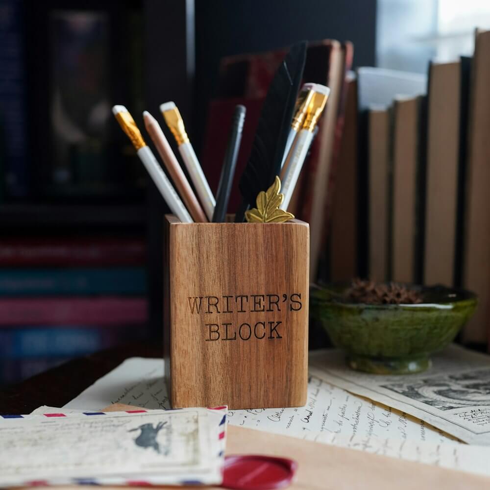 Writer's Block Pen Holder is a wooden pen cup with "writer's block" written on the side.