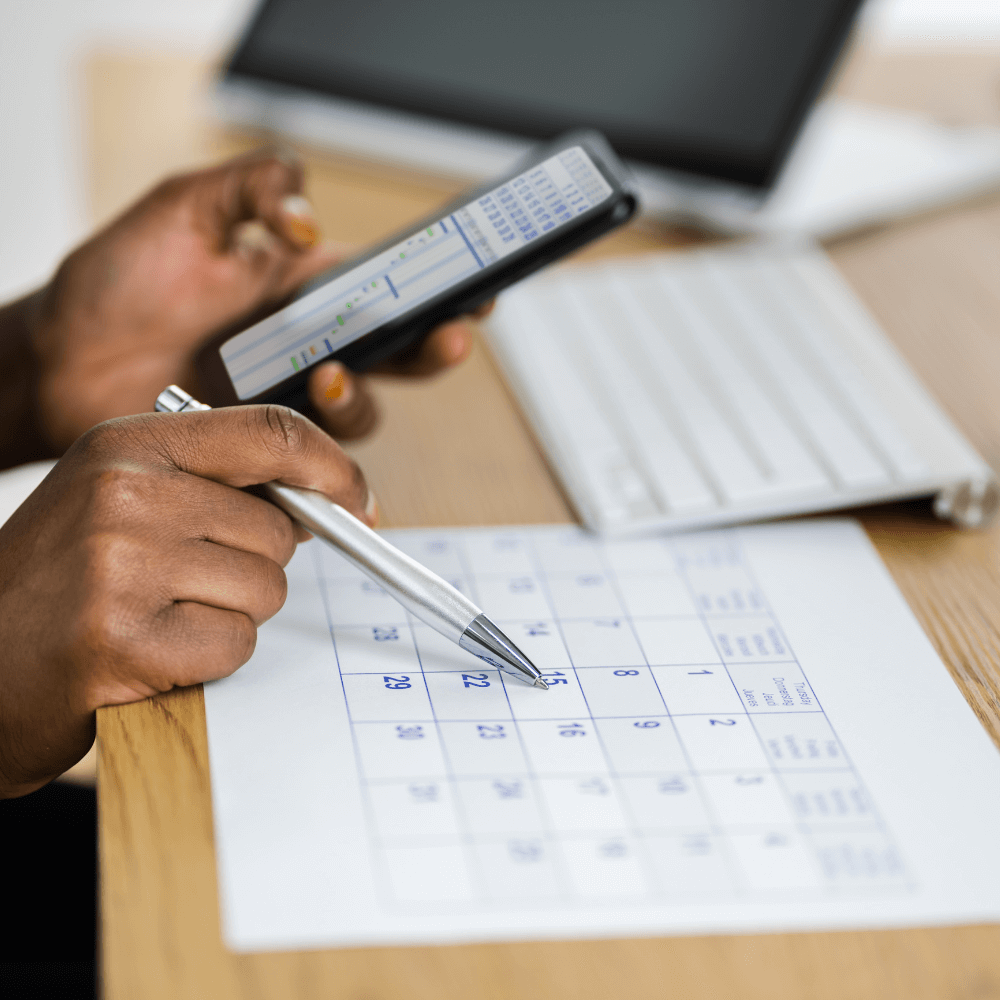 a person's hands holding a phone in their left and writing on a calendar with their right