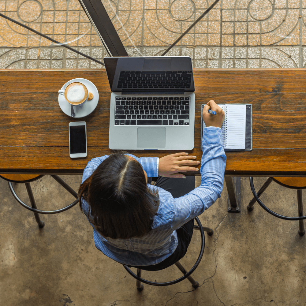 a woman sitting at a coffee shop with a laptop, a coffee, a phone, and a notebook in front of her