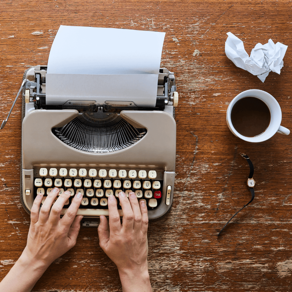 hands typing at a typewriter with a crumpled paper and a coffee next to them
